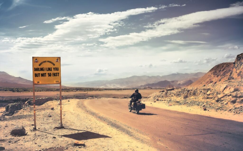 A motorcyclist rides through the vast, barren deserts of Ladakh with a humorous road sign in view.